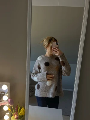 Grå stickad tröja med stora prickar - Mysig grå stickad tröja med stora färgglada prickar i brunt, vitt och beige. Tröjan har en avslappnad passform, rund halsringning och långa ärmar. Perfekt för dig som gillar en lekfull och trendig stil.