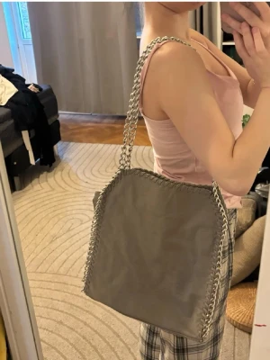 Grå axelväska med kedjedetaljer - Snygg grå axelväska med silverfärgade kedjor längs kanterna och som axelrem. Väskan har en mjuk, fyrkantig form och är tillverkad i ett slätt material som liknar veganskt läder. Perfekt för dig som gillar trendiga accessoarer med lite edge. Lite sliten men syns knappt