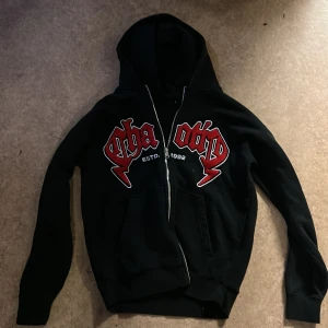 Svart hoodie med röd brodyr från FSBN - Svart hoodie från FSBN i relaxed fit med dragkedja och huva. Framsidan har stora röda broderade bokstäver och texten 'ESTD. 1992' i vitt. Mjuk bomullskänsla, kängurufickor och långa ärmar. Perfekt för en streetwear-look.