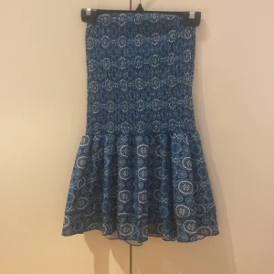 Blå kortklänning med mönster - Supersöt blå kortklänning med smock upptill och volangig kjol. Klänningen har ett detaljerat vitt och blått mönster som ger en bohemisk vibe. Perfekt för varma dagar och enkel att styla med sneakers eller sandaler.