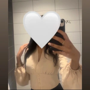 Beige blus med v-ringning och knappar - Säljer en beige blus med v-ringning och dekorativa knappar framtill. Blusen har långa puffärmar med resår vid handlederna och en figurnära passform upptill. Perfekt för dig som gillar stilrena och trendiga plagg.