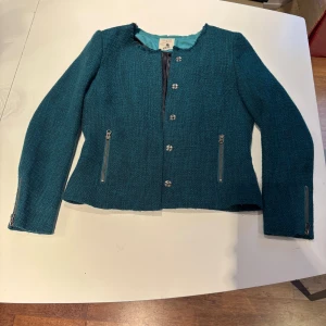 Turkos kavajjacka från Jackpot - Snygg turkos kavajjacka från Jackpot med grov textur, rund hals och råa kanter. Jackan har silverfärgade dragkedjor vid fickor och ärmslut samt dekorativa knappar framtill. Fodrad insida i satin och diskret röd logotyp på ryggen. Perfekt för dig som gillar stilrena detaljer.