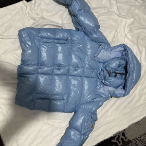 Ljusblå Moncler pufferjacka med huva - Säljer en snygg ljusblå pufferjacka från Moncler med glansig finish och huva. Jackan har flera dragkedjefickor, klassisk Moncler-logga på ärmen och coolt serietryck på insidan. Perfekt för kalla dagar när du vill sticka ut med stil.