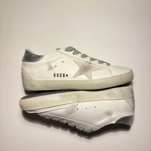  Golden Goose skor  - Snygga vita sneakers från Golden Goose med grå snörning, silverfärgad häl och ikonisk stjärndetalj i mocka på sidan. Skorna har en platt sula och är tillverkade i skinn med detaljer i mocka. Perfekta för en cool och avslappnad stil. De är helt nya 