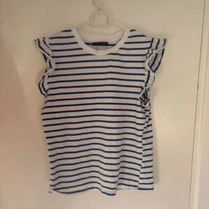 Randig volangtopp från SHEIN XS - Säljer en vit och blå randig topp från SHEIN i storlek XS. Toppen har volangdetaljer vid axlarna och är ärmlös. Perfekt för en fräsch och somrig look. Tillverkad i mjuk bomull för extra komfort.