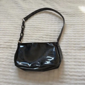 Svart axelväska med kedjedetalj - Snygg svart axelväska i glansigt syntetmaterial med cool kedjedetalj på axelremmen. Väskan har en stilren och enkel design med mjuka former och passar perfekt för dig som gillar trendiga accessoarer.