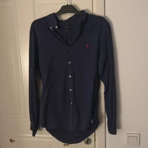 Blå skjorta från Ralph Lauren slim fit - Snygg marinblå skjorta från Ralph Lauren i slim fit med klassisk krage och knappar framtill. Tillverkad i lätt bomullstwill och har den ikoniska röda broderade loggan på bröstet. Perfekt för dig som gillar stilrena och tidlösa plagg.