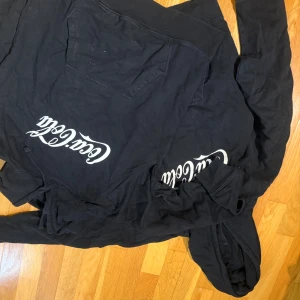 Svart hoodie från Coca-Cola - Svart hoodie med stor vit Coca-Cola-logga på framsidan. Tröjan har huva, lång ärm och en klassisk känguruficka. Perfekt för dig som gillar streetwear och vill sticka ut med en ikonisk logga. Säljer 2 för 200