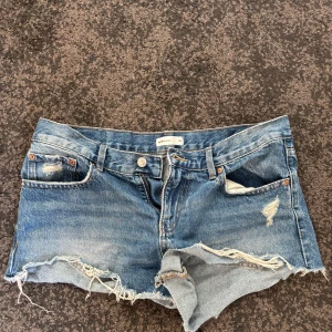 Låg midjade blå jeansshorts med slitningar - Säljer ett par blå jeansshorts från Gina tricot med råa kanter och slitna detaljer. Klassisk femficksmodell med knapp och dragkedja. Perfekta för varma dagar och en avslappnad look. 💞💞
