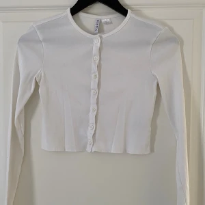 Vit croppad topp från H&M Divided - Säljer en vit croppad topp från H&M Divided med rund hals och långa ärmar. Toppen har knappar framtill och är tillverkad i mjuk bomull. Enkel och stilren design som är lätt att matcha med olika outfits.