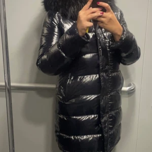 Svart  lång pufferjacka med päls - Säljer en svart, glansig pufferjacka med lång passform och stor fluffig päls runt huvan. Jackan har dragkedja framtill och är perfekt för kalla dagar. Den har en snygg, modern look och håller dig varm hela vintern.