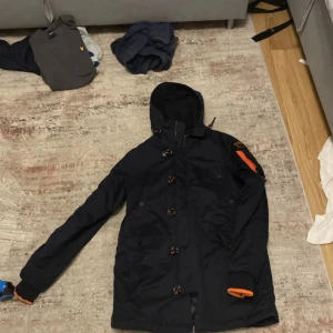 Svart parkajacka Superdry XL - Svart parkajacka från Superdry i storlek XL. Jackan har huva, stora fickor, orange detaljer och dragkedja på ärmen. Tillverkad i microfibrer och polyester, med coola knappar och broderad logga på ärmen. Perfekt för kalla dagar och streetstyle.
