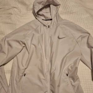 Ljusgrå Nike vindjacka med huva - Nike vindjacka i ljusgrått med huva och hel dragkedja framtill. Jackan har två fickor med dragkedja, diskret Nike-logga på bröstet och ventilerande panel på ryggen. Perfekt för löpning eller utomhusaktiviteter. Snygg och sportig design.
