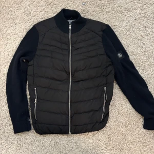 Svart quiltad pufferjacka med stickade ärmar - Svart pufferjacka från L’agency med quiltad front och rygg i syntetmaterial samt stickade långärmade ärmar.