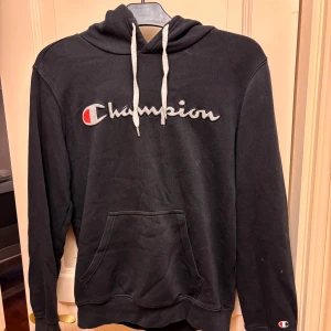 Svart Champion hoodie Stl S - Svart hoodie från Champion i storlek S med broderad logga i vitt och rött på bröstet. Tröjan har huva med vita snören och en stor magficka framtill. Mjuk och skön. Fin!