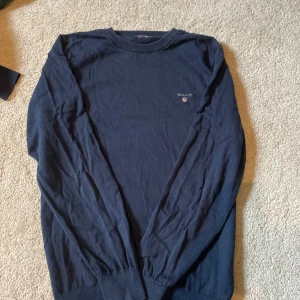 Mörkblå bomullströja från GANT - Snygg mörkblå tröja från GANT i 100% bomull. Klassisk rund halsringning och diskret broderad logga på bröstet. Långärmad modell med ribbade muddar för en clean och stilren look. Perfekt för dig som gillar enkel och tidlös stil.