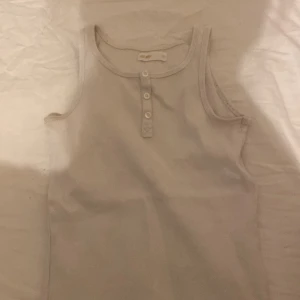 Beige ribbad linnetopp 157 New York - Säljer en beige ribbad linnetopp från 157 New York i storlek 146. Toppen är ärmlös med rund halsringning och har tre knappar framtill som snygg detalj. Perfekt för en enkel och clean look.
