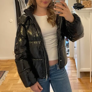 Svart glansig pufferjacka från Nelly - Säljer en svart glansig pufferjacka med stor huva och dragkedja framtill. Jackan är quiltad och har en oversized passform med långa ärmar. Knappt använd🩷