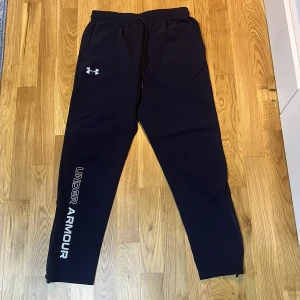 Mörkblå träningsbyxor Under Armour L - Snygga mörkblå träningsbyxor från Under Armour med vit logga på låret och stort tryck längs benet. Byxorna har snörning i midjan, elastisk resår och dragkedja vid benslut. Perfekta för gymmet eller chill hemma. 
