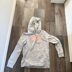 Ljusgrå hoodie från Vans - Säljer en ljusgrå hoodie från Vans med rosa logga framtill. Tröjan har huva med snörning och en stor magficka. Perfekt för dig som gillar en avslappnad och cool stil.