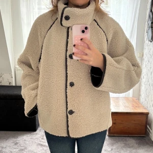Beige teddyjacka med vändbar design - Supermysig vändbar teddyjacka i beige och mörkbrun med stora knappar och bred krage. Ena sidan är fluffig teddy i beige med svarta detaljer, andra sidan är slät mörkbrun med teddyfoder. Jackan har en loose passform och långa ärmar – perfekt för kalla dagar.
