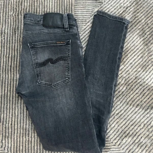 Svarta jeans från Nudie Jeans - Säljer ett par svarta skinny jeans från Nudie Jeans i storlek W30 L32. Jeansen har klassisk femficksdesign, svarta sömmar och Nudie-loggan på bakfickan. Snygg tvättad look och stretchigt material för skön passform.