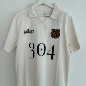 Vit FC Barcelona Lamine Yamal tröja - Säljer en vit fotbollströja med FC Barcelona-emblem och trycket '304' på framsidan. Tröjan har klassisk krage, korta ärmar och är tillverkad i ett lätt, sportigt material. Baksidan har en stor grafisk illustration och texten 'Lamine Yamal'.