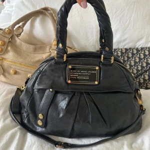 Svart Q Classic väska med gulddetaljer - Snygg och praktisk Marc by Marc Jacobs Classic Q väska i mjukt svart pebble-läder med guldfärgade detaljer. Stor svart Marc by Marc Jacobs bricka. Flera fack som gör det till en väldigt smidig vardagsväska. Dubbla handtag och långt band följer med 🤍  Inuti finns dragkedjefack och öppna fack för enkel organisering. En populär modell ur Classic Q-serien och en eftertraktad vintage-väska.  Kan hämtas i Nacka eller skickas. Skriv för mer info och bilder 🤍