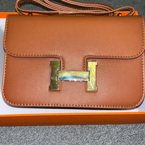 Dam väska 💼  - Säljer en brun axelväska med klassiskt H-spänne i guldmetall. Väskan är tillverkad i exklusivt skinn och har en stilren, rektangulär form med justerbar axelrem. Insidan har flera fack för smart organisering. Perfekt för dig som vill ha en ikonisk och lyxig accessoar.