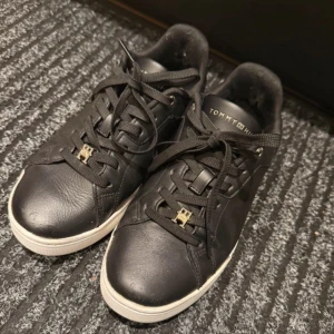 Svarta sneakers Tommy Hilfiger  - Snygga svarta sneakers från Tommy Hilfiger i skinn med vita sulor och gulddetaljer på snörningen och sidan. Klassisk låg modell med rund tå och svarta skosnören. Märkeslogga på plösen och sidan. Perfekta för en stilren och trendig look. Lite slitet i skorn som man ser på bilden, men de ändrar verkligen inte på passformen och dom funkar perfekt och som helt nya skor! 
