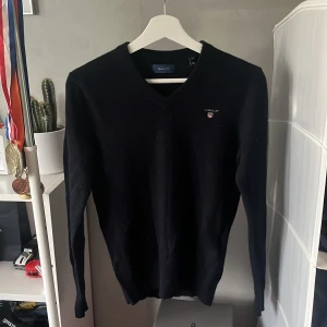 Mörkblå v-ringad tröja från GANT - Mörkblå v-ringad tröja från GANT med broderad logga på bröstet. Tröjan har lång ärm och ribbade muddar vid hals, ärmslut och nederkant. Perfekt för dig som gillar klassisk och stilren look.