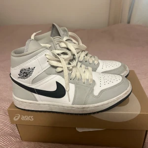Nike Air Jordan 1 Mid vit/grå - Säljer ett par Nike Air Jordan 1 Mid i vitt och ljusgrått med svart swoosh. Klassisk high-top siluett med snörning och perforerad tå. Skorna har ikoniska Air Jordan-loggan på sidan och är tillverkade i skinn och syntet. Perfekta för dig som gillar streetwear och sneakers med retrovibe. Använda men är fortfarande i ett bra skick.
