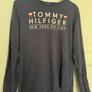 Blå långärmad tröja Tommy Hilfiger - Mörkblå långärmad tröja från Tommy Hilfiger med stort tryck framtill och klassisk logga. Tillverkad i ekologisk bomull, rund halsringning och relaxed fit. Perfekt för dig som gillar stilren och enkel design med en touch av street.