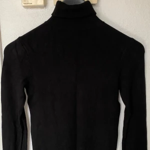 Svart polotröja med pärlknappar - Snygg svart polotröja med lång ärm och coola pärlknappar på ärmsluten. Tröjan är stickad och har en klassisk polokrage som ger en stilren vibe. Perfekt för dig som gillar detaljer och vill ha något som sticker ut lite extra.