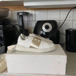 Valentino Garavani sneakers vit/guld - Snygga vita sneakers från Valentino Garavani med bred guldfärgad detalj över mitten. Skorna har klassisk snörning, platt sula och är tillverkade i skinn. Perfekta för dig som vill ha en clean men lyxig look med en touch av guld.