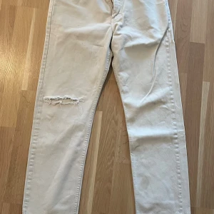 Vita Levi's jeans med slitning - Säljer ett par vita Levi's jeans med snygg slitning på ena benet. Jeansen har klassisk rak passform och är tillverkade i bomull. Perfekta för dig som gillar en clean look med lite edge. Finns lite fläckar som kan gå bort i tvätten!