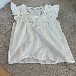 Vit blus med spets och volang - Säljer en söt vit blus från VILA med spetsdetaljer och broderade mönster upptill. 