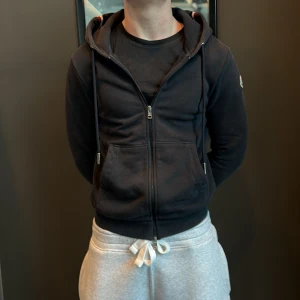 Navy blue hoodie från Moncler med dragkedja - Navy blue hoodie från Moncler med hel dragkedja, huva och snörning. Tröjan har Moncler-logga på ärmen och ribbade muddar med rödvita detaljer. Tillverkad i mjuk bomull, perfekt för chill dagar. Klassisk och stilren design med känguruficka framtill. (Passar dig som är 168-183)
