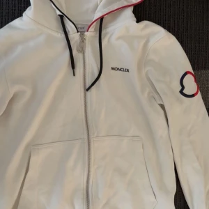 Vit Moncler hoodie med logga, S - Vit hoodie från Moncler med dragkedja, svart snörning och broderad logga på bröstet samt ärmen. Stora fickor framtill och röd detalj på luvans kant. Perfekt för dig som gillar stilrena och exklusiva plagg.