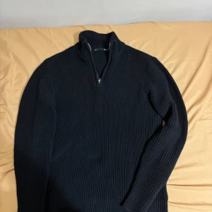 Svart stickad half zip-tröja 157 - Svart ribbstickad tröja från 157 med hög krage och half zip-dragkedja. Tröjan är långärmad och har en klassisk, stilren look som passar perfekt till lager-på-lager. Materialet är grovstickat och ger en mysig känsla. Nytt skick storlek M 