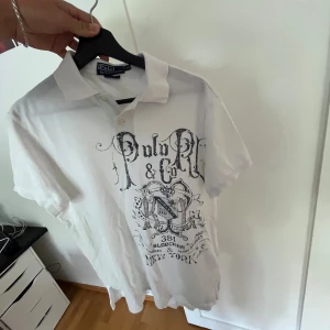Vit Polo Ralph Lauren t-shirt med tryck - Vit Piké från Polo Ralph Lauren med svart/grått tryck framtill och klassisk krage med knappar. Modellen är custom fit och har korta ärmar. Perfekt för dig som gillar street och stilren design. 