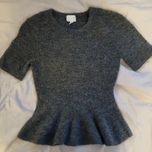 Grå stickad topp från H&M - Mysig grå stickad topp från H&M med korta ärmar och markerad midja som ger en snygg peplum-effekt. Perfekt för lager-på-lager och passar till både jeans och kjol. Ribbad struktur och rund halsringning.
