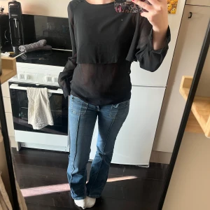 Svart blus med transparenta ärmar från UNIQ - Svart blus från UNIQ i storlek XS med transparenta, långa ärmar och ett lager över bröstet. Blusen har rund halsringning och knäppning bak i nacken. Tunn och luftig känsla, perfekt för en stilren look.