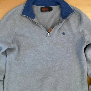 Grå merinoulltröja från Morris - Snygg grå tröja från Morris i 100% merinoull med blå krage och halv dragkedja med läderdetalj. Broderad logga på bröstet och ribbstickad krage ger en clean och stilren look. Perfekt för lager-på-lager under kyliga dagar.