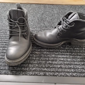 Svarta kängor från Ecco i skinn - Svarta kängor från Ecco i slätt skinn med snörning och rund tå. Insidan är fodrad med mjukt material för extra komfort. Grov sula som ger bra grepp. Perfekta för kyligare dagar och enkel att matcha med olika Storlek 38.