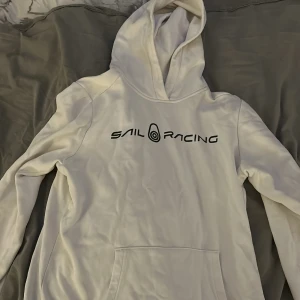 Vit hoodie från Sail Racing - Vit hoodie från Sail Racing med svart logga tryckt över bröstet. Klassisk huva och stor magficka framtill. Perfekt för dig som gillar en enkel och stilren look. passar till många outfits.