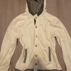 Beige hoodiejacka med grå detaljer - Säljer en beige hoodiejacka med grå detaljer på huva, fickor och muddar. Jackan har knäppning med knappar framtill och är tillverkad i mjuk bomull. Perfekt för lager-på-lager och chill vardagsstil.