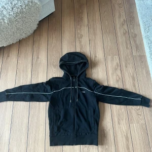 Svart Hugo boss zip hoodie  - Svart hoodie från HUGO BOSS med vit piping längs ärmar och bröst.Tillverkad i mjuk bomull, perfekt för en chill och stilren look. Använd få gånger, köpt ifrån butiken Hugo boss och säljs ej längre