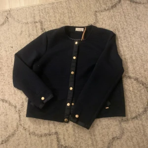 Mörkblå cardigan jacka Holly & Whyte - Snygg mörkblå cardigan jacka från Holly & Whyte by Lindex med guldfärgade knappar framtill och vid ärmslut. Jackan har rund halsringning och är långärmad, perfekt för att ge din outfit en stilren touch.