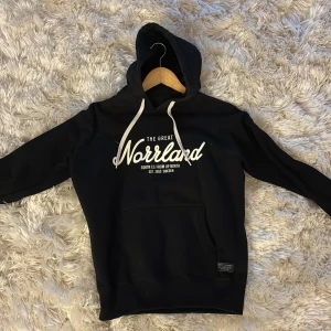 Svart hoodie från SQRTN Company - Svart hoodie från SQRTN Company med vit text 'The Great Norrland' på bröstet och vit snörning i huvan. Stor känguruficka framtill och en liten hjortron-detalj på ärmen. Mjuk bomullskänsla och ribbade muddar. Perfekt för dig som gillar norrländsk stil.